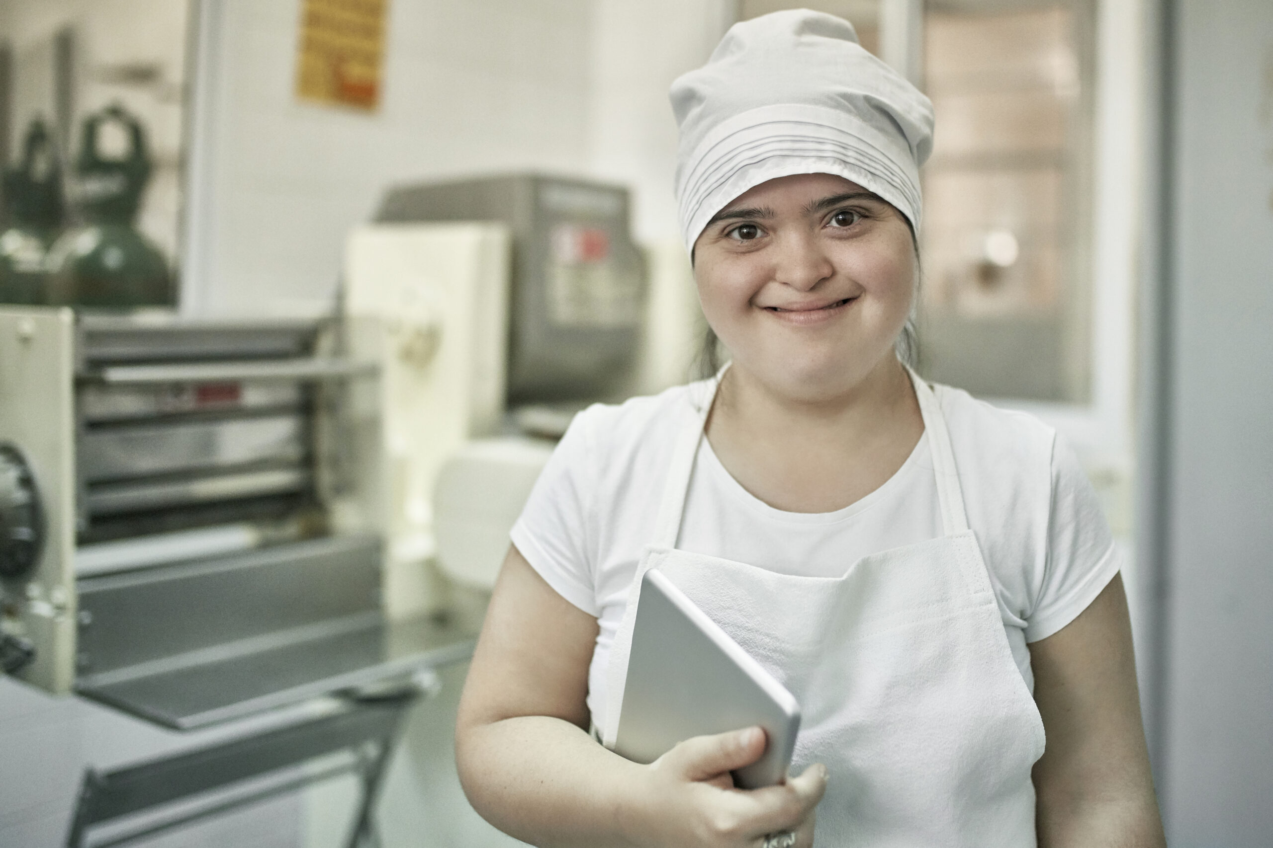 Close-up of smiling female worker with Down Syndrome holding digital tablet and wearing apron and hat while working in Argentine pasta factory.