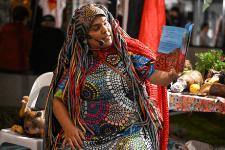 A woman in colourful dress holds up a book as she reads to children out of shot